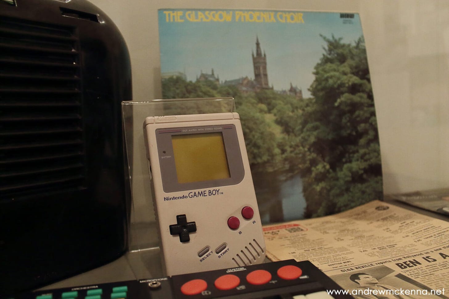 A Nintendo Gameboy and a 12 inch LP that has a cover photo of Glasgow university sprie through the trees.