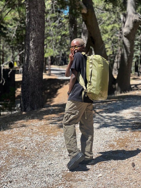 photo of Oumou Sylla with blonde buzzcut wearing glasses, a black tee, green cargos, green backpack, holding up a peace sign in the woods