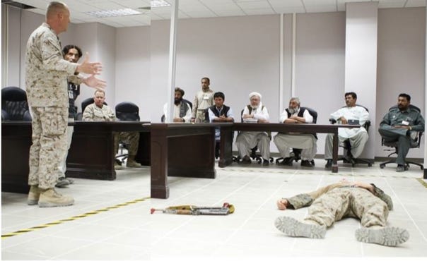 The image shows a military training in a conference room in Helmand, Afghanistan in 2011. A U.S. Marine in camouflage uniform is standing and addressing a group of seated individuals; a mix of military personnel and local officials or leaders. The seated men, dressed in traditional clothing and police or military uniforms, are attentively listening. In the foreground, a soldier is lying on the floor as part of a demonstration, with a rifle placed nearby.  