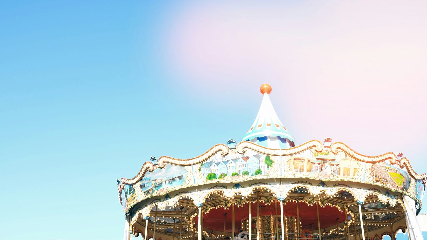 Pastel photograph of the top third of a lovely carousel, its spire reaching into a pastel blue sky with fluffy clouds