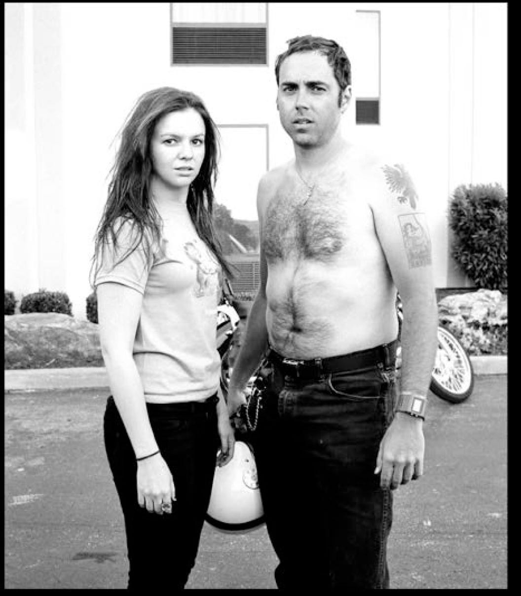 Amber and Derrick standing in a parking lot. The both are turned in, facing each other slightly, but looking toward camera. Derrick is shirtless. They both wear odd/tired expressions. Behind them, the motorcycle can be seen partially. 
