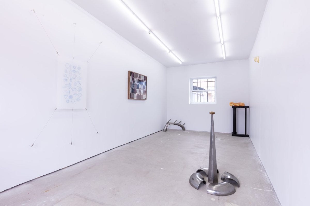 Installation photo shows a white-walled gallery with a silver floor sculpture in the foreground, two wall pieces and two sculptures in the background