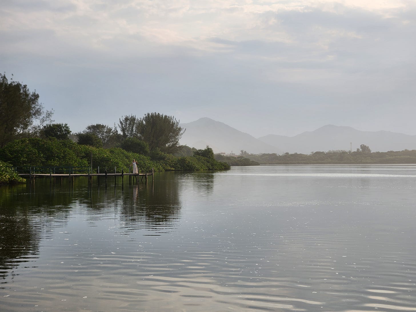 over a river, a dock emerges from trees and a woman in a dress stands there, under the cloudy sky.