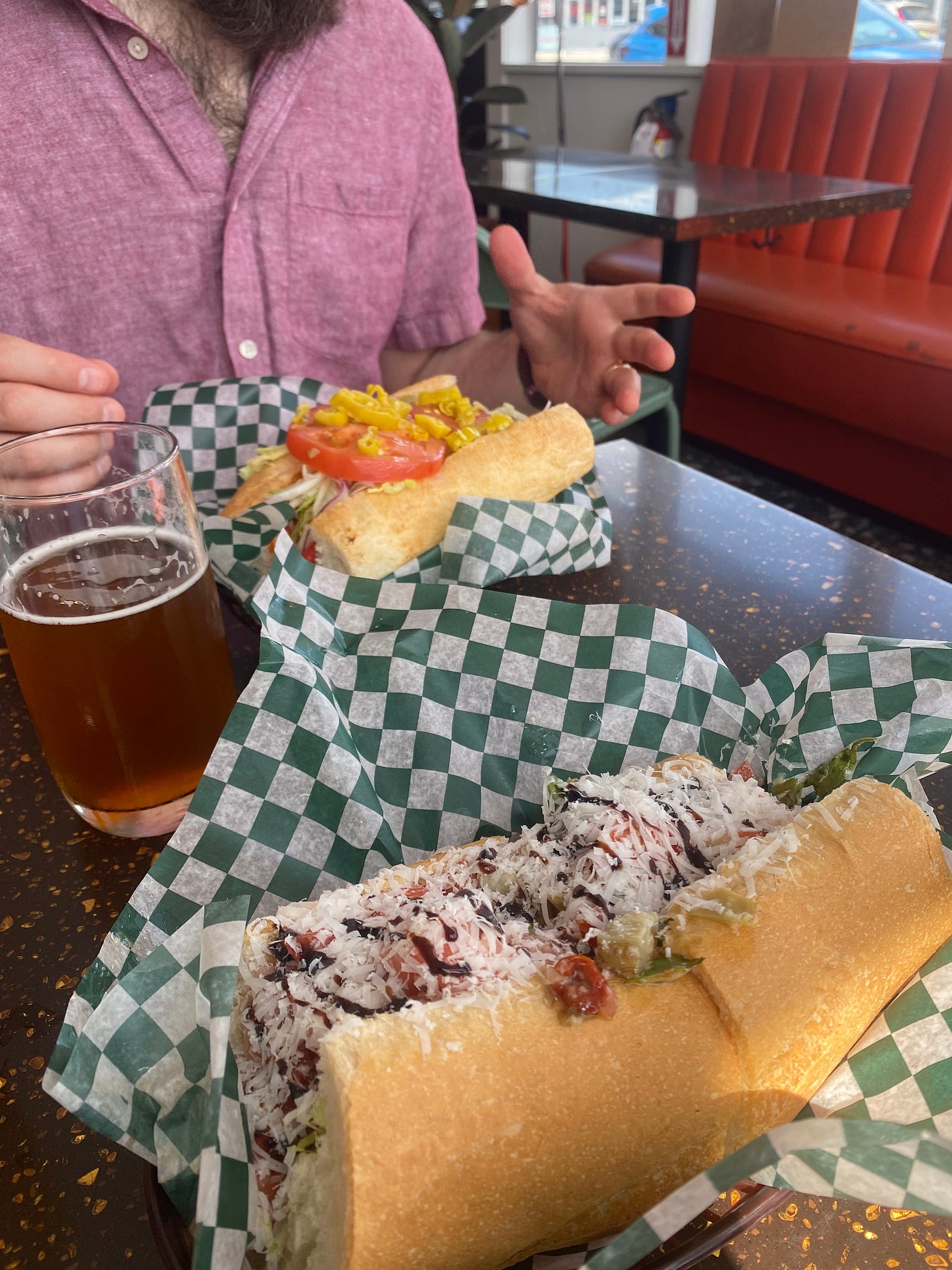 Two big subs in baskets lined with checkered deli paper. The nearest one has lots of parmesan and a little balsamic drizzle, and the other has tomato and pepperoncini. There's a glass of beer on the table between them. In the background, Jeff is wearing a red chambray shirt and holding his hands on either side of the sandwich, like he's not sure how to pick it up.