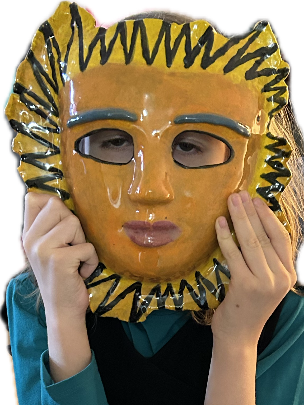 A child holds a large, glossy, handmade ceramic mask in front of their face. The mask is painted in warm yellow and orange tones with bold black zigzag patterns around the edges, resembling a sunburst. The mask has blue painted eyebrows, cut-out eye holes, and sculpted lips. The child's eyes peek through the openings, and their hands gently hold the sides of the mask. They are wearing a dark vest over a teal long-sleeve shirt.