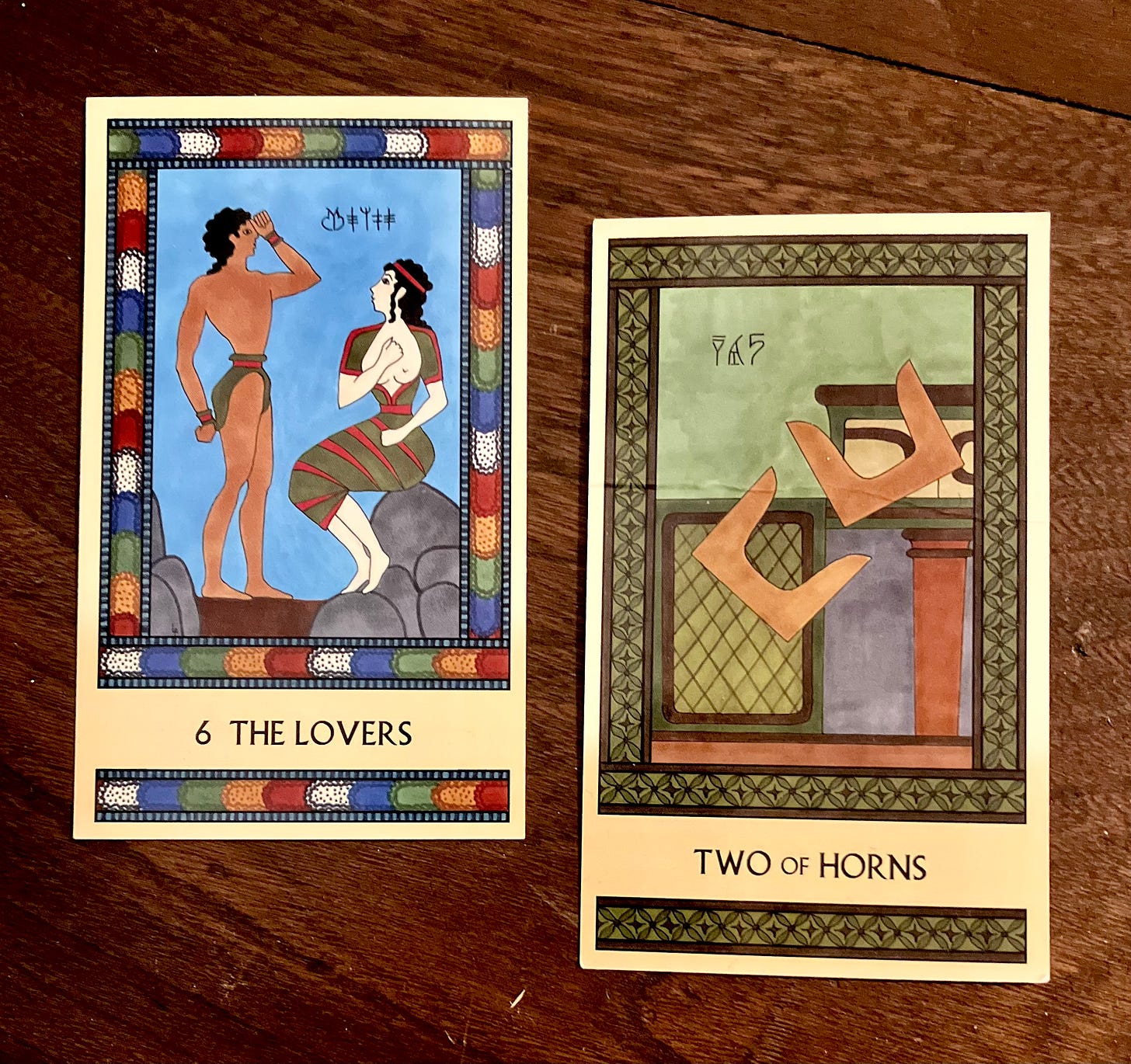 Two Minoan Tarot cards lie on a dark brown wood surface. The Lovers has a multicolored border. It depicts a Minoan man standing, facing right, holding his fist up to his forehead. He is facing a Minoan woman who is seated, facing left, holding her fist to her chest. The Two of Horns is painted in shades of deep green and brown. It shows two pairs of Minoan sacred horns in midair as they tumble off a shrine.