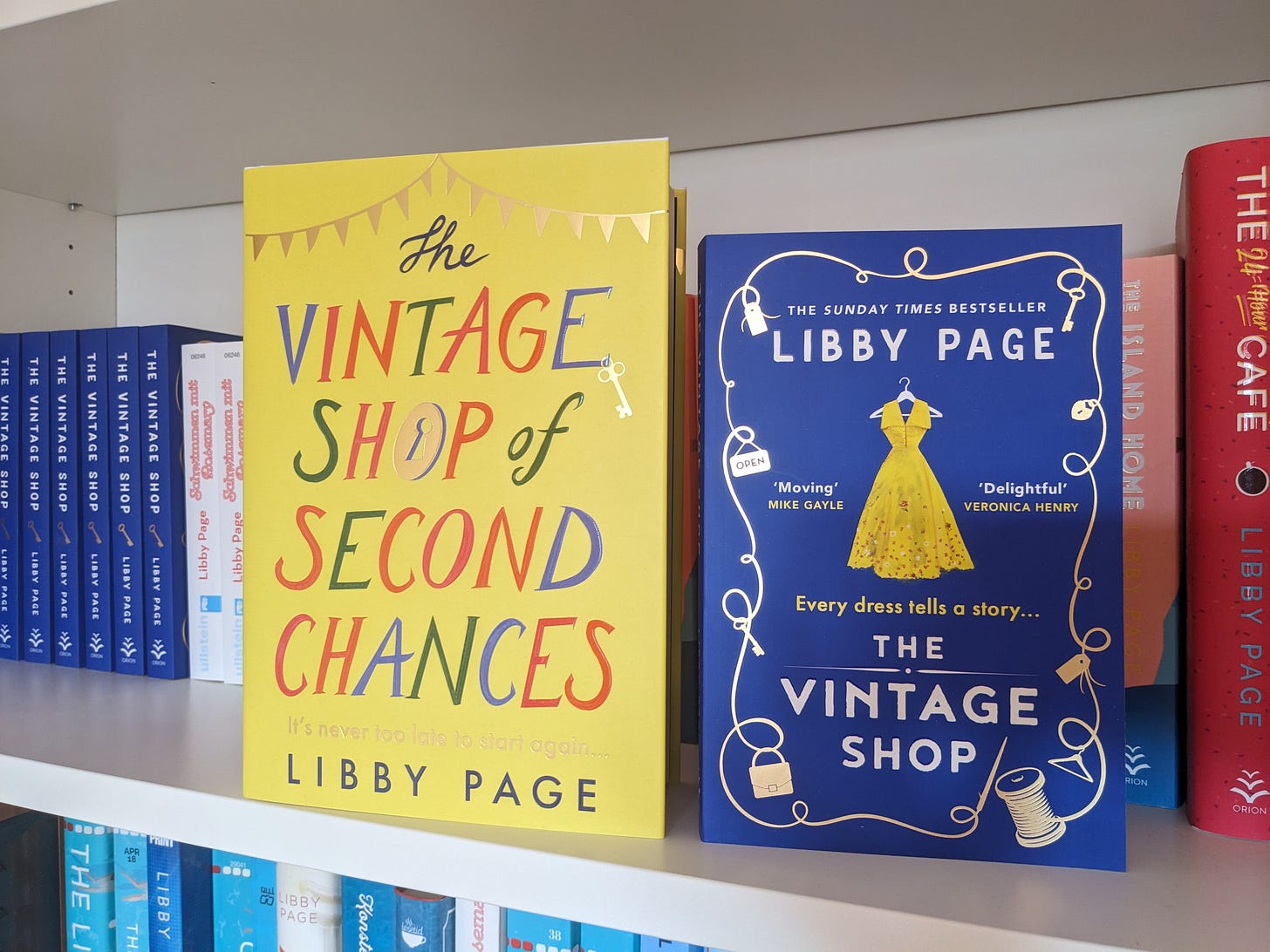 The hardback covers of The Vintage Shop by Libby Page next to the paperback edition on a shelf. The hardback is bright yellow and text-based. The paperback is blue and has a yellow dress in the middle and a reel of thread creating a border with sewing paraphernalia. 