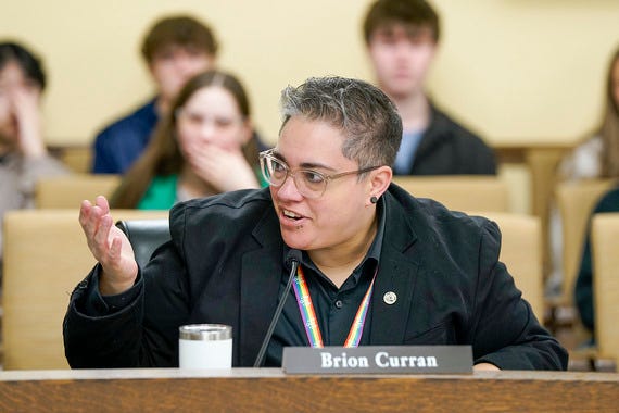 A queer person with short salt and pepper hair is speaking into a microphone wearing a rainbow lanyard.
