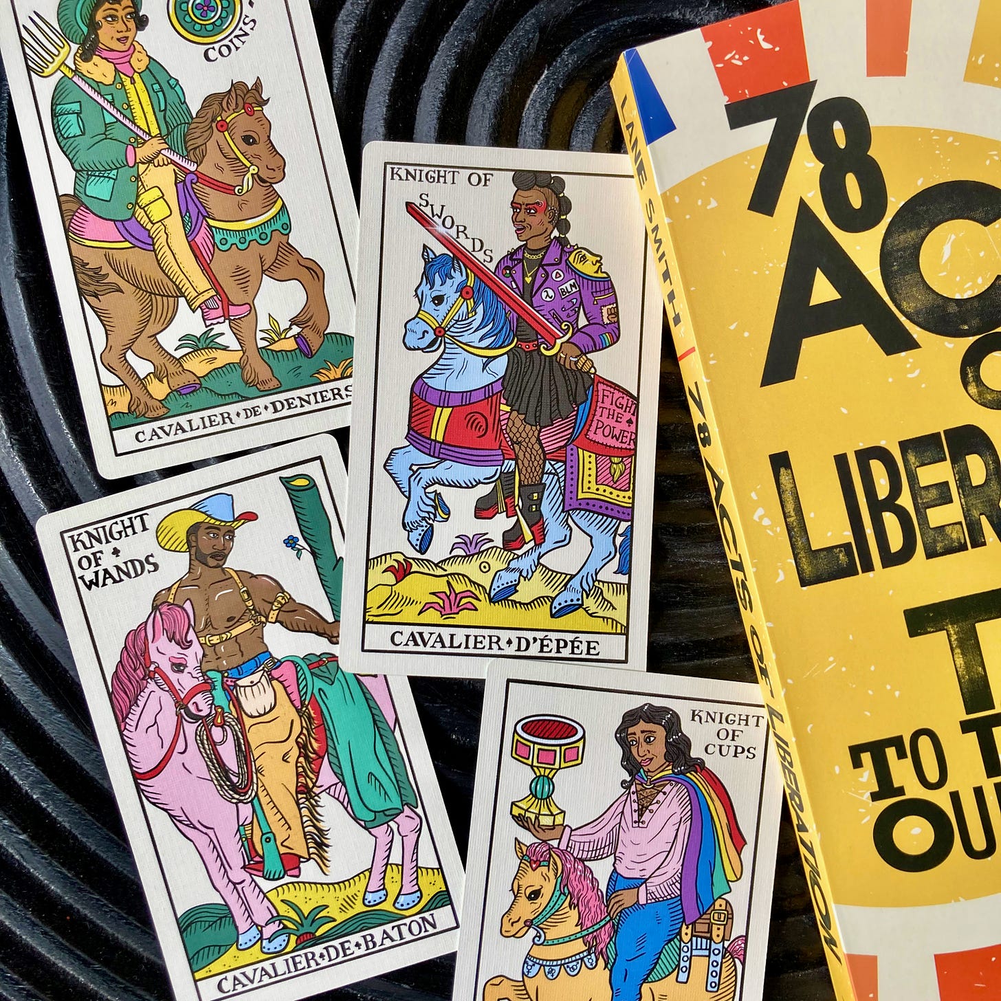 Four brightly colored tarot cards on a black table next to a copy of 78 Acts of Liberation. The cards are the four Knights and the focus is on the Knight of Swords, which has an illustration of a Black person riding a light blue horse. The person wears big boots, fishnet stockings, and a punk-style jacket with patches including a lambda, "BLM," and the Palestinian flag.