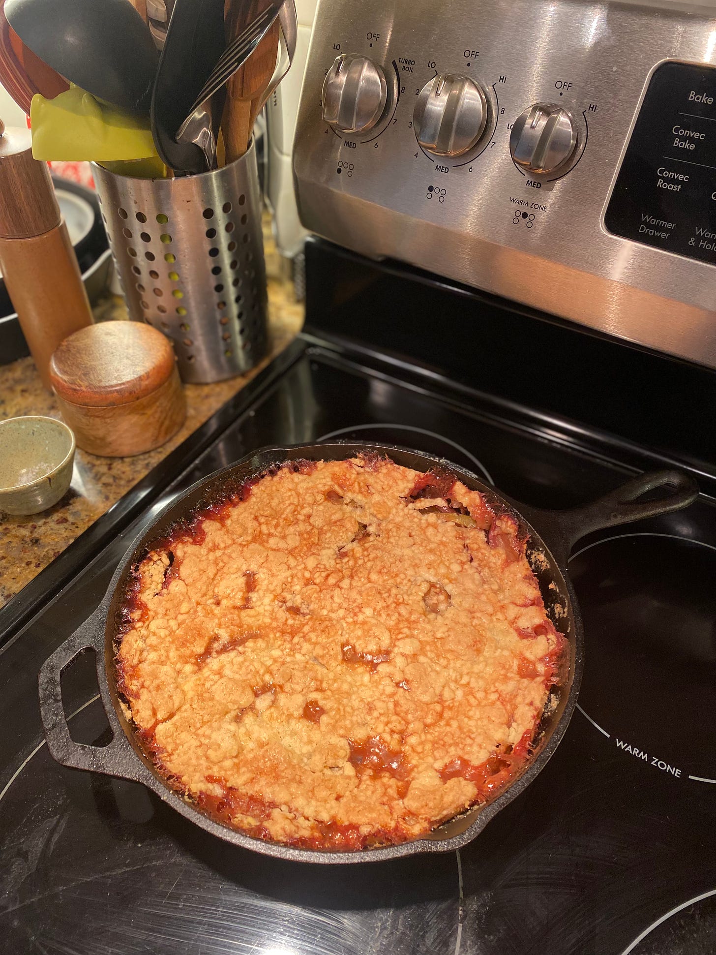 A cast iron pan on top of a stove. The pan is full of the apple cobbler described above, with pinkish areas at the edges where the fruit has caramelized, and a crackly-looking golden top.