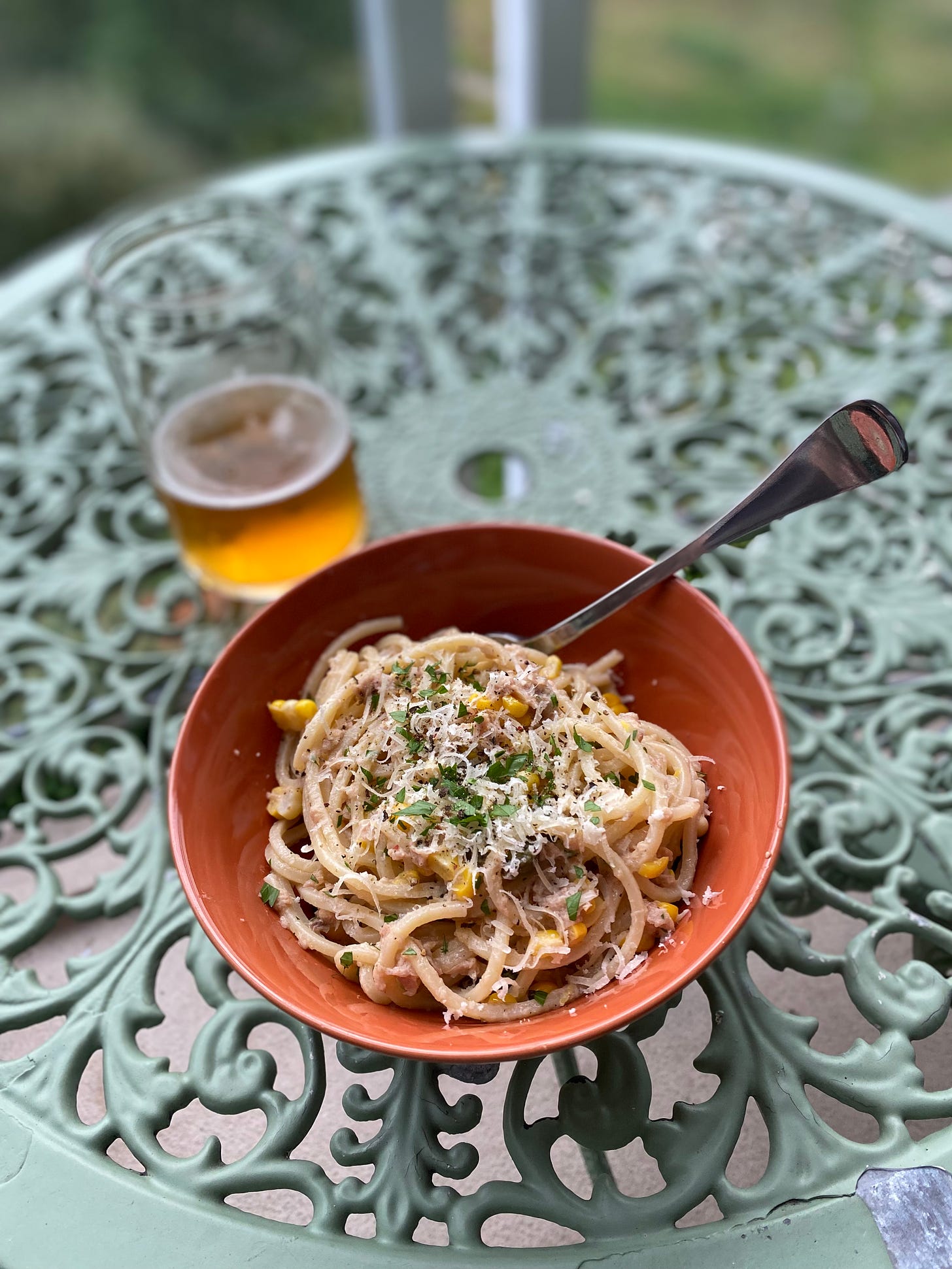 On an outdoor café table, an orange bowl of the pasta described above, topped with grated parmesan and chopped parsley. A mostly-empty glass of beer is in the background.