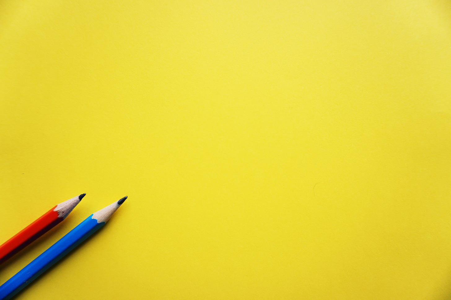 Sfondo uniforme giallo. Dall'angolo in basso a sinistra spuntano in posizione obliqua due matite, una rossa e una azzurra.