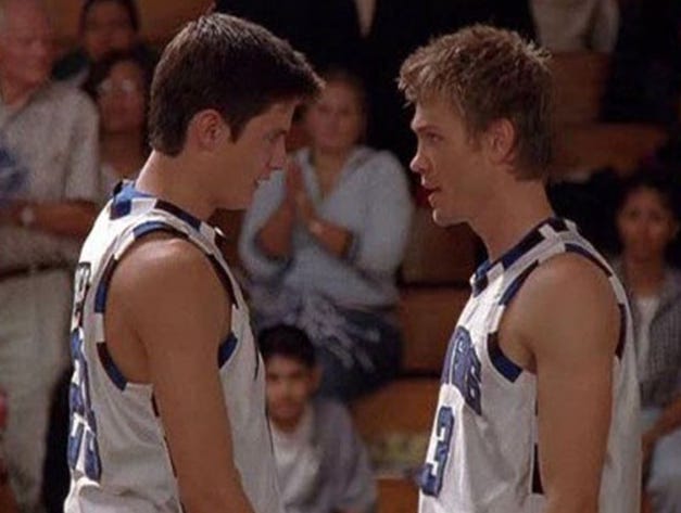 Lucas and Nathan from One Tree Hill, wearing basketball uniforms and staring each other down in the court as the crowd watches