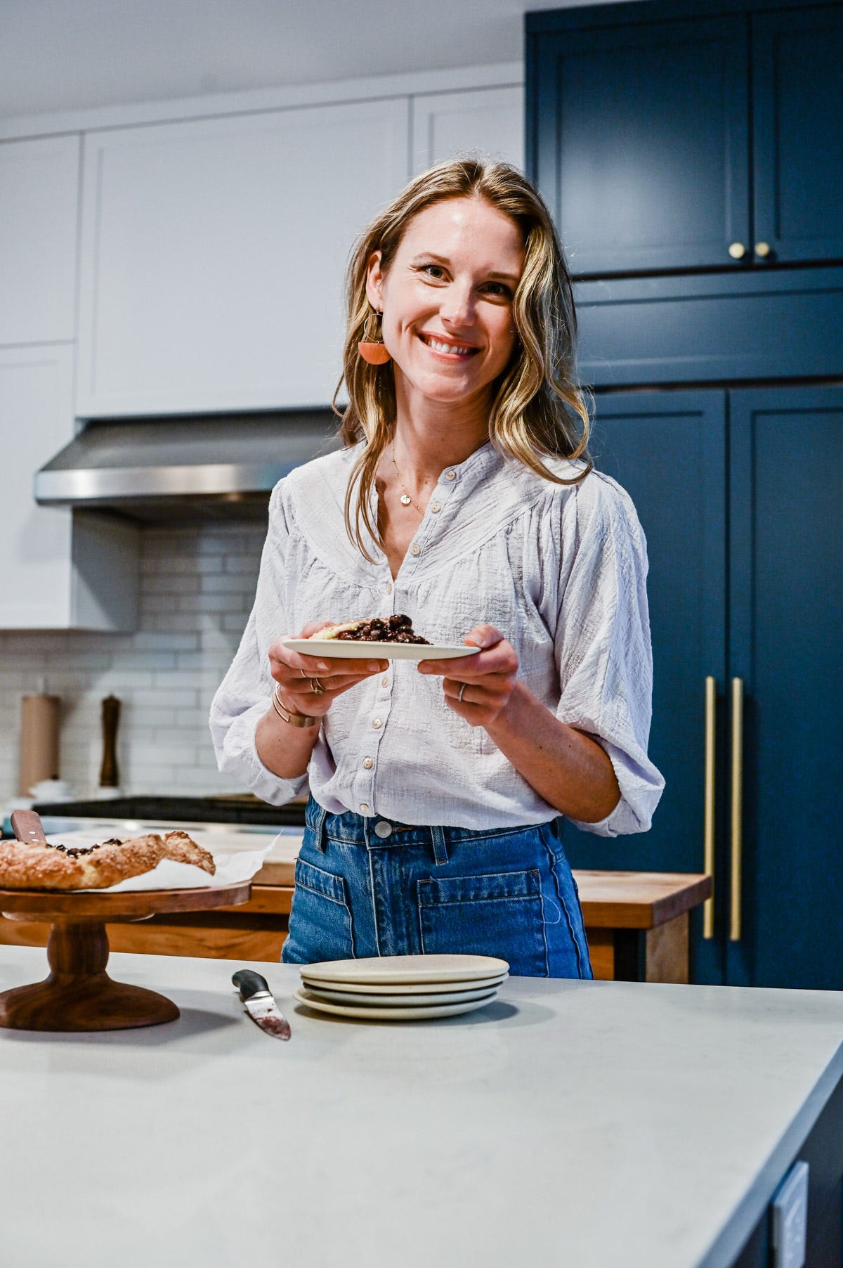 Nicki, wearing a white top and blue jeans, in her kitchen holding a plate of food.