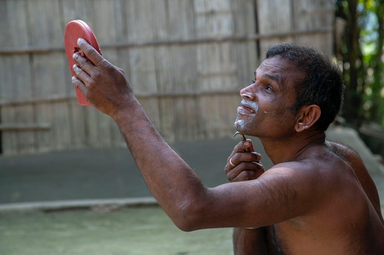 Gobinda, a middle-aged DeafBlind Indian man with short black hair is sitting on the ground without a garment on his torso. He is holding up a mirror with a red plastic frame in his left hand. A razor in his right hand runs over his chin as he looks into the mirror and shaves off his facial hair. Shaving cream covers parts of his chin, upper lip region and left ear. 