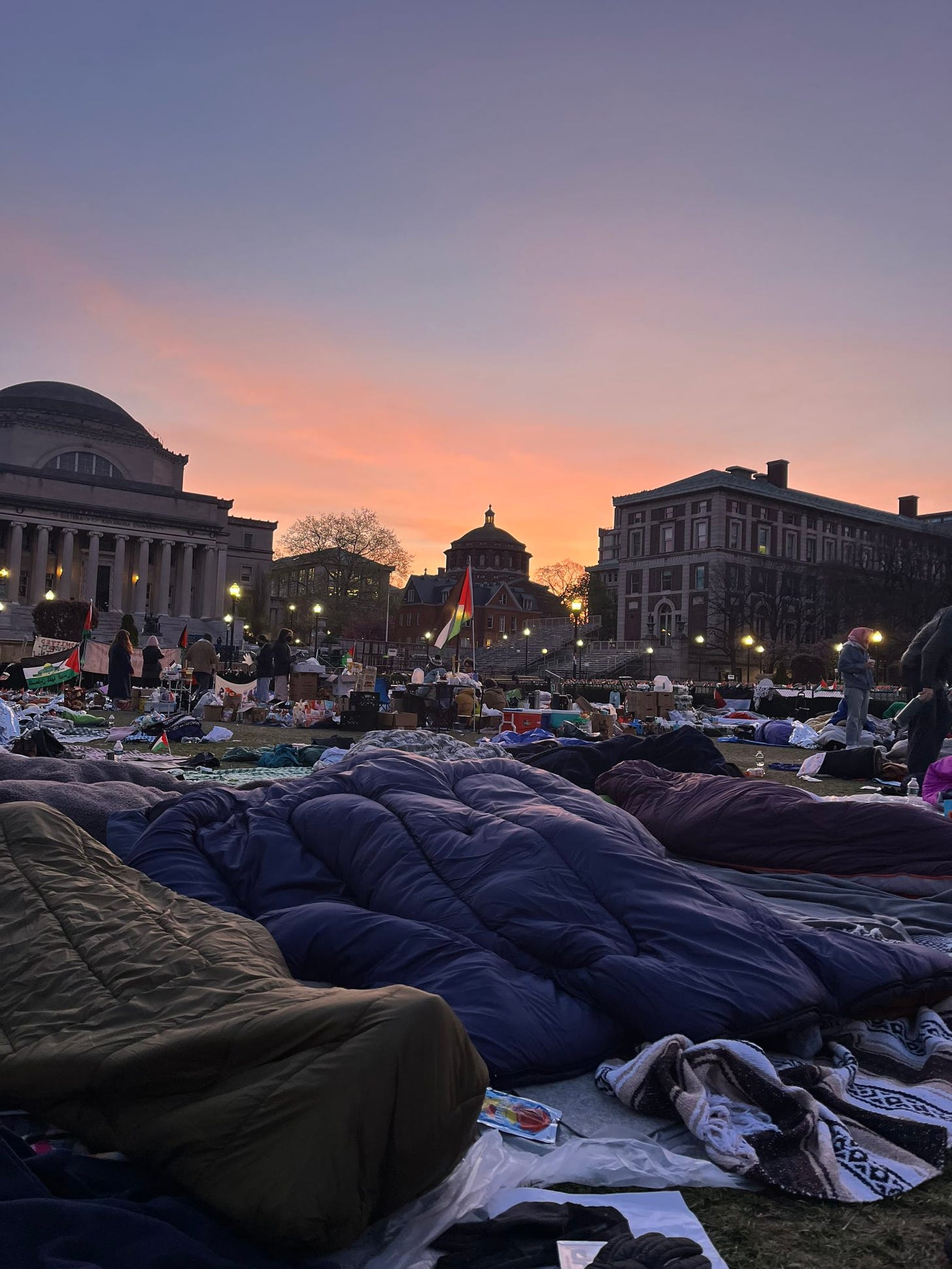 The sun rises over Columbia University. The campus streetlights are still on while students slowly rise out of sleeping bags across the lawn. The Palestinian flag can be seen in several places across frame. Stations, coolers, and supplies are in the background. This photograph has been taken from a tweet (linked in the photo and in the caption)