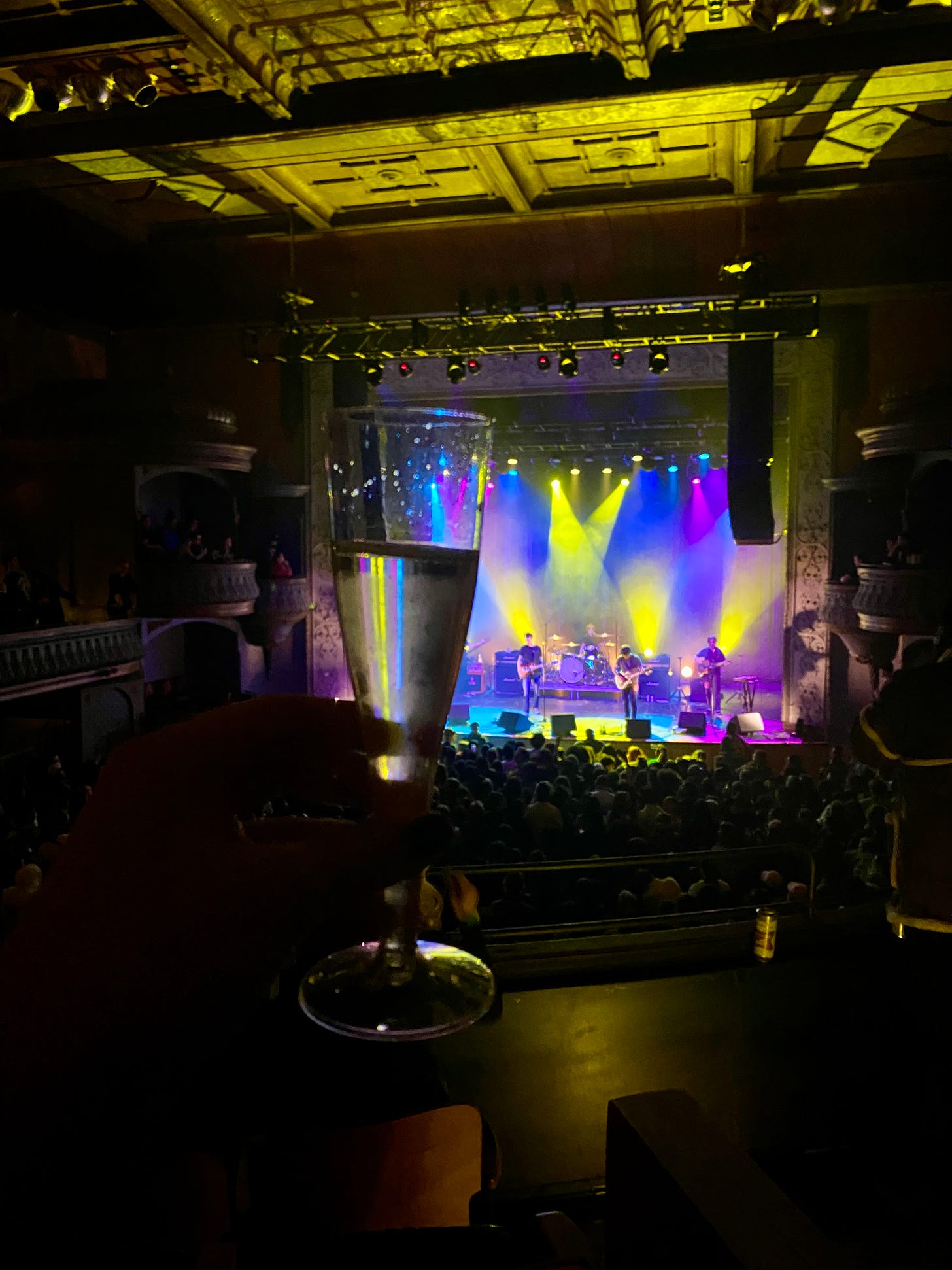A photo of a hand holding a glass of champagne up in the air. In the background is an audience standing in front of a stage with the band Joyce Manor on it. Yellow, blue, and purple lights shine down on the stage while the rest of the theater is dark.