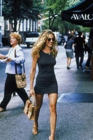 This may contain: a woman is walking down the street in a short black dress and gold heels with her handbag This may contain: a woman is walking down the street in a short black dress and gold heels with her handbag
