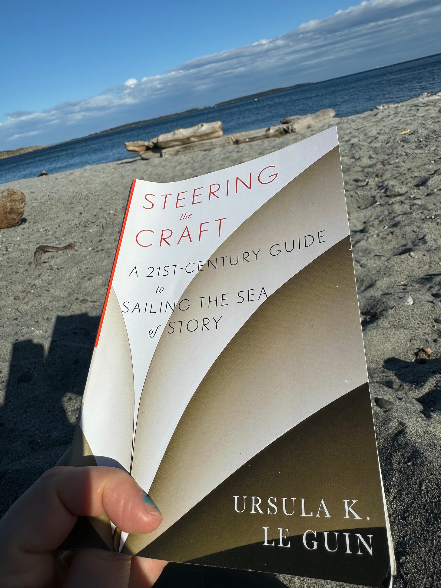 A hand holds up a copy of Steering the Craft: A 21st-Century Guide to Sailing the Sea of Story by Ursula K. Le Guin against the backdrop of a sandy beach and ocean view.