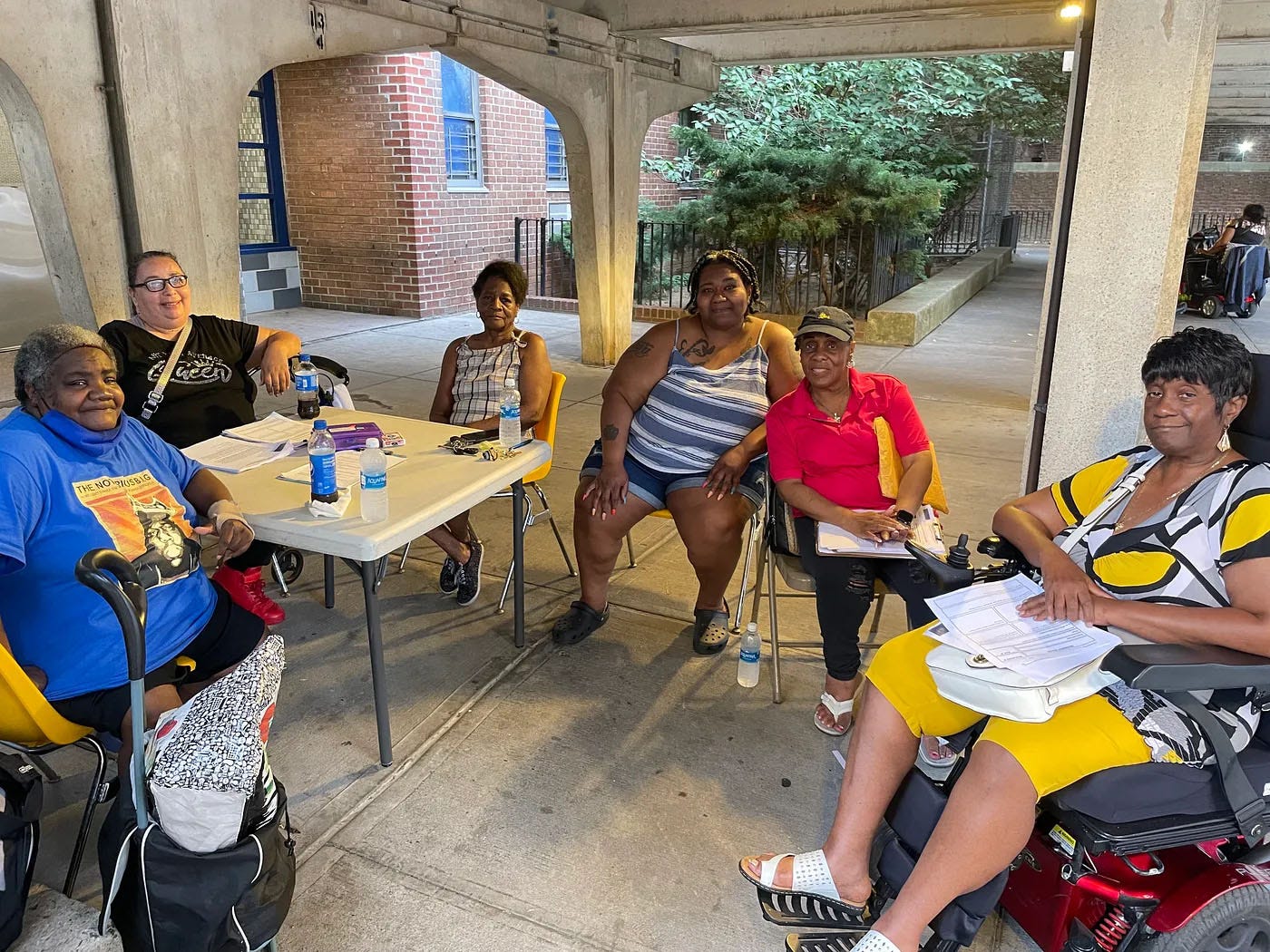 Six Black women sit around a card table under a concrete portico, with a brick building and foliage behind them.