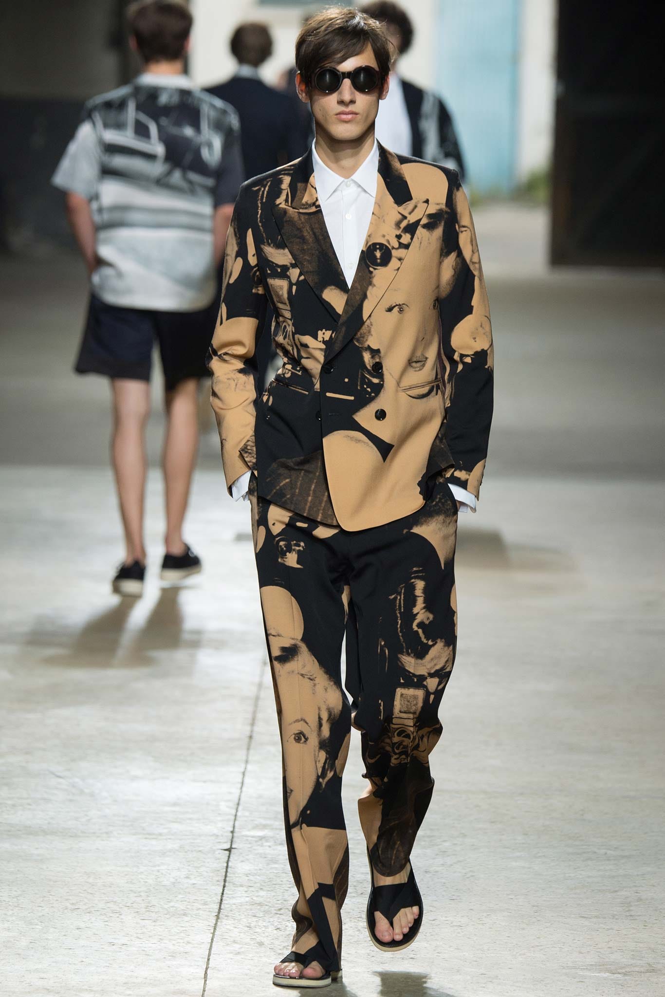 A model on a fashion catwalk, who is a white man with dark brown hair, wearing sunglasses, and a double breasted suit with an all over orange and black print featuring the oversize image of Marilyn Monroe, and black sandals with a chunky sole. A model on a fashion catwalk, who is a white man with dark brown hair, wearing sunglasses, and a double breasted suit with an all over orange and black print featuring the oversize image of Marilyn Monroe, and black sandals with a chunky sole.