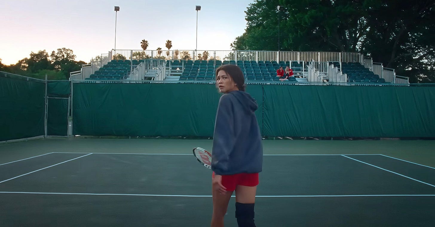 Movie still from Challengers. Zendaya stands in an empty tennis court, holding a racket and looking over her shoulder.