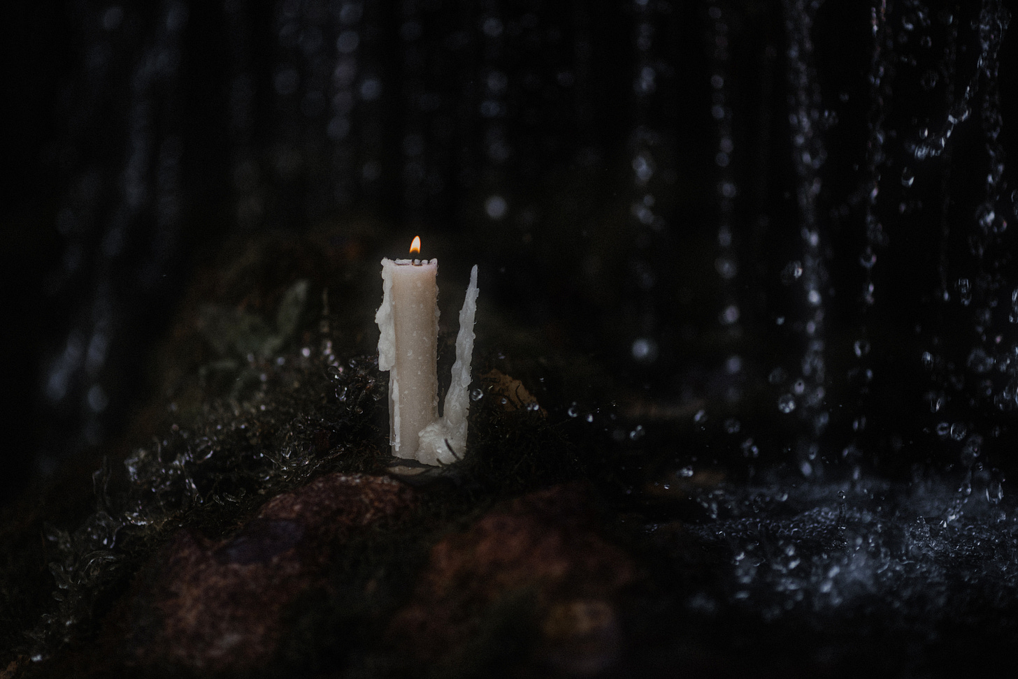 Lit white candle on a rock with dark background as rain falls around it
