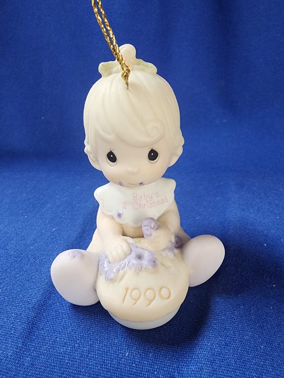 A ceramic figurine of a little girl seated, eating blueberry pie with her hands. Her bib reads Baby's 1st Christmas. 1990 is written on the pie.