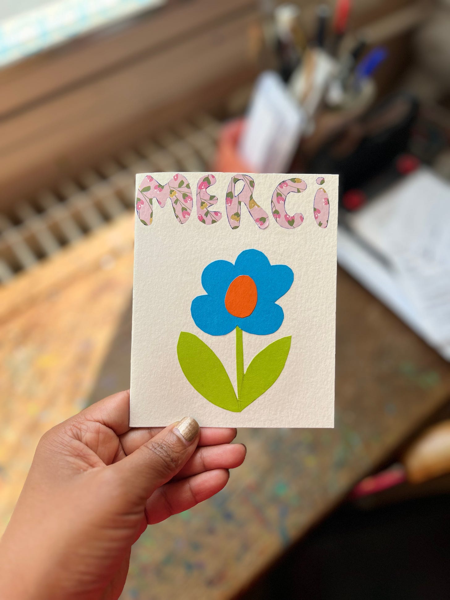A white card with a collage of the word merci and a blue flower with green leaves
