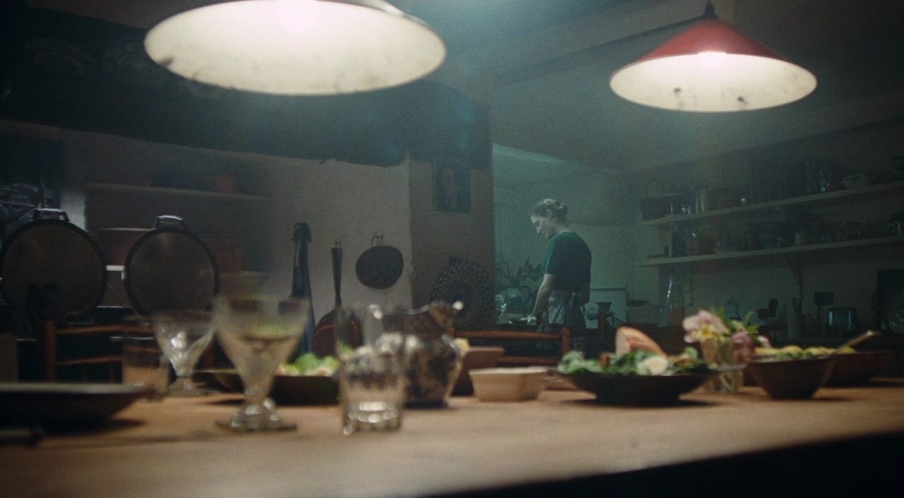 a white woman wearing a dark green shirt stands over the stove in the corner of a country house kitchen with glasses, salads, and a loaf of bread on a set table in the foreground.
