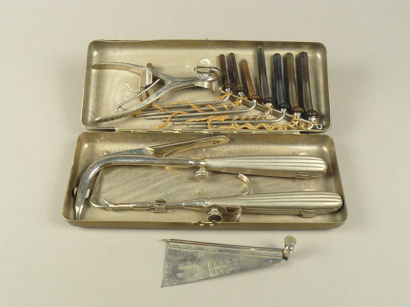 Small, flat, metal case with various sugical instruments including a small saw blade and hollow metal tubes with rubber handles. Small, flat, metal case with various sugical instruments including a small saw blade and hollow metal tubes with rubber handles.