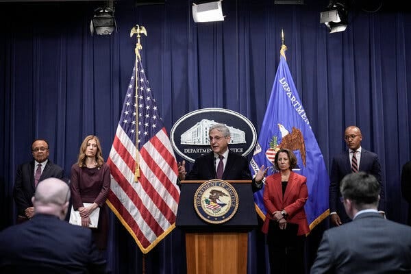 Attorney General Merrick B. Garland, flanked by people on both sides, stands behind a lectern that is displaying the seal of the Justice Department.