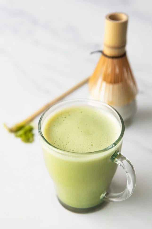A close up image of a matcha latte, with a traditional matcha whisk and scoop in the background