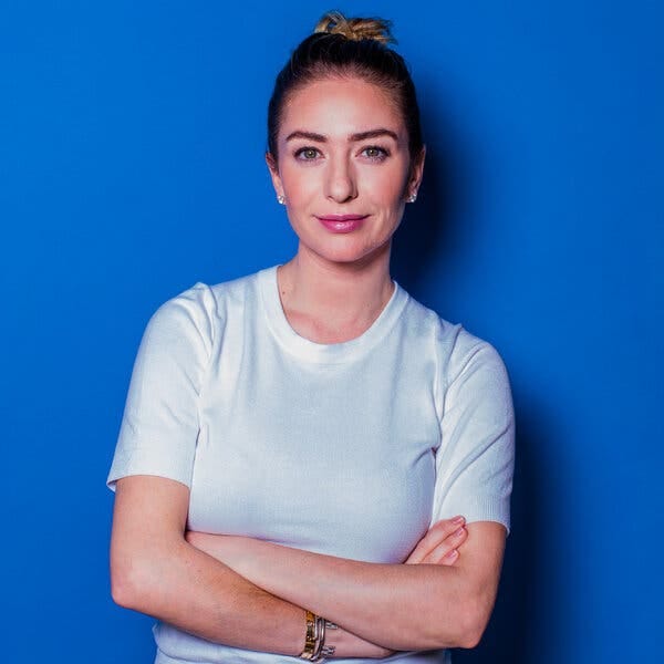 A portrait of a smiling woman posing in a white T-shirt against a blue background with her arms folded in front of her.