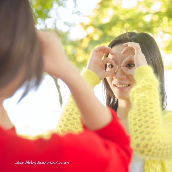 Two women look at each other making goggles with their hands. One woman has brown hair and a yellow sweater. The other has brown hair and a red sweater.