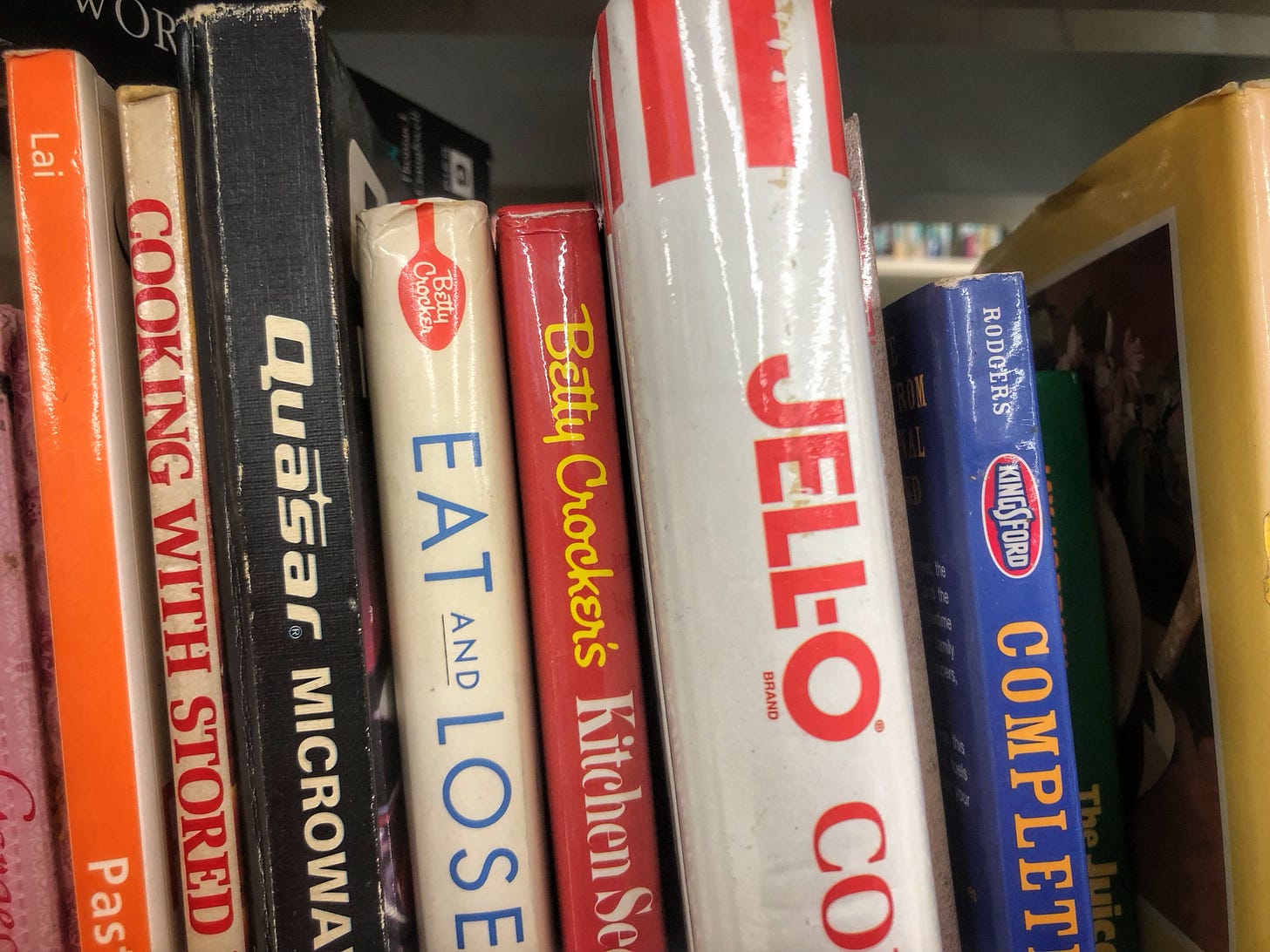 The spines of cookbooks published by Quasar Microwaves, Betty Crocker, Jell-O, and Kingsford Charcoal on the shelf in a thrift store