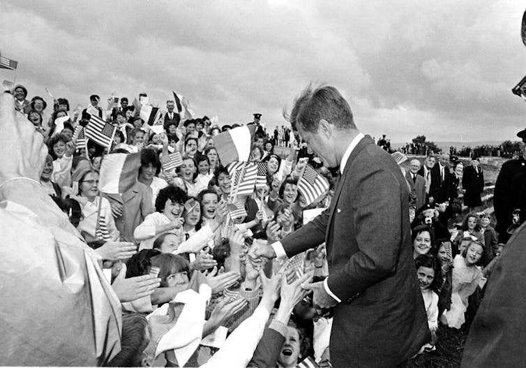 A man in a suit shakes hands with flag-waving wellwishers.