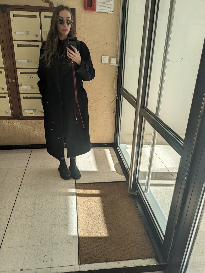 Sara stood in a hallway with sunglasses and a long black coat