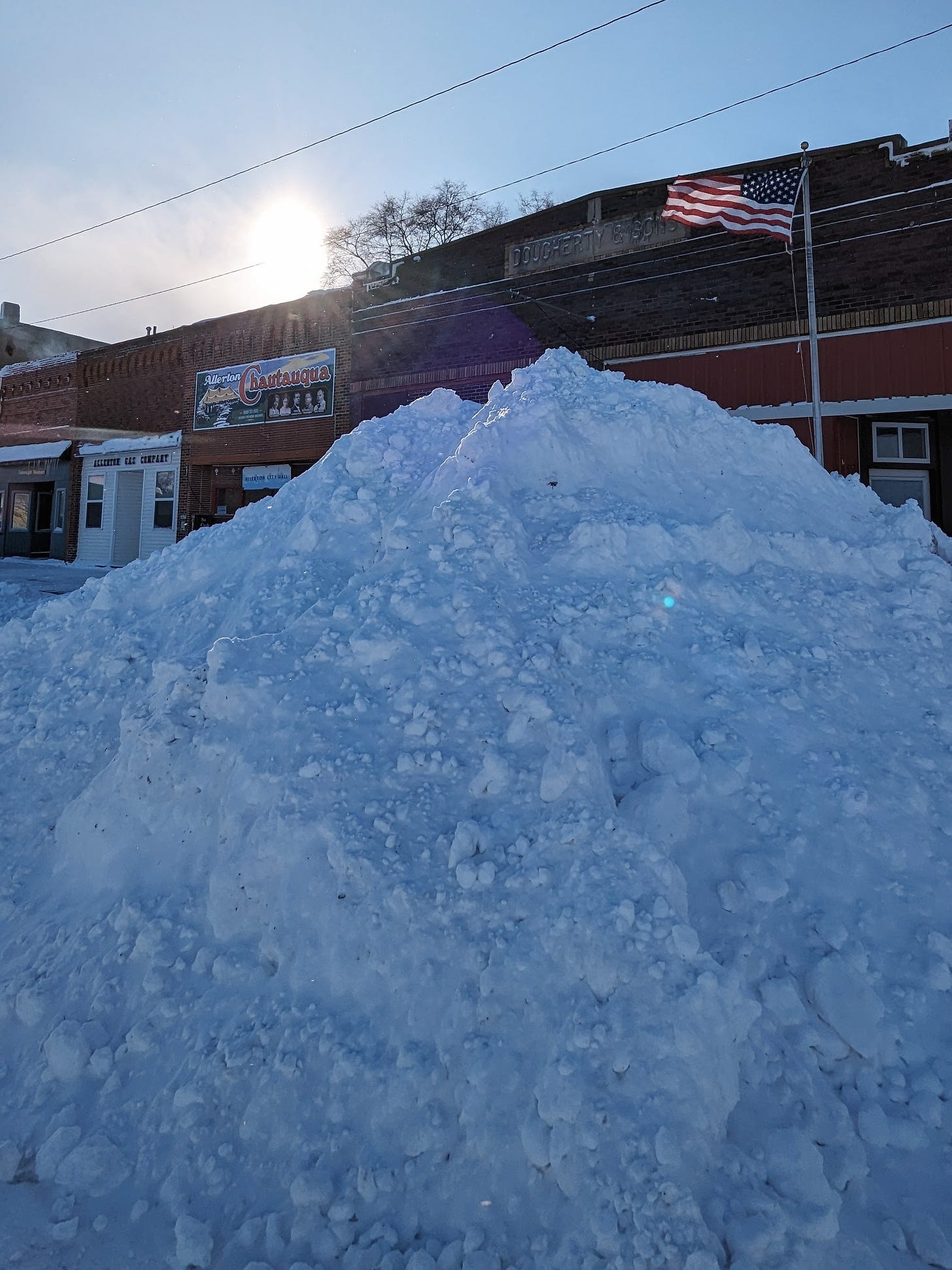 A large pile of snow in front of several brick buildings and an American flag, with sun setting behind it