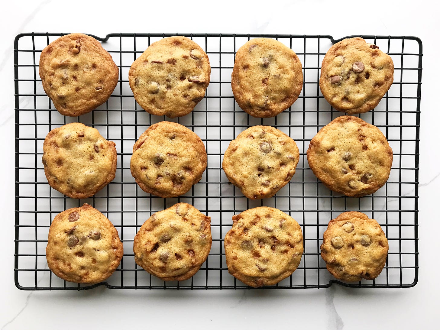 One dozen Chocolate Chip Cookie with Toffee and Pecans on a rectangular black wire cooling rack. Each cookie is different, some with more chocolate chips, others with an extra nut or two, some have little craters where the toffee bits have melted. Their edges are a golden brown while the centres are paler.