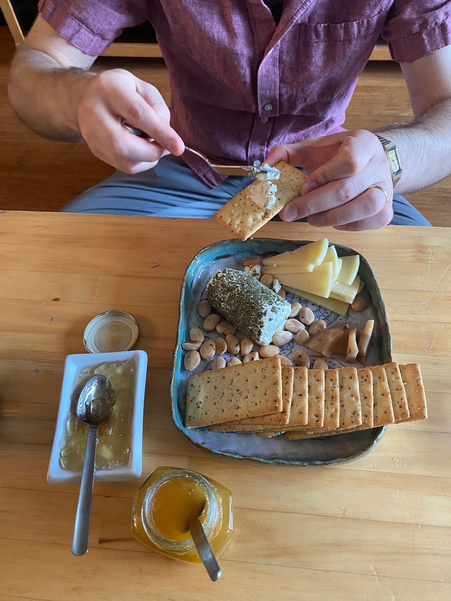 Jeff is sitting on the floor in front of the coffee table, arranging a cracker with cheese above a small spread of two cheese, rectangular poppyseed crackers, a few pickled beets, marcona almonds, and a couple of condiments.