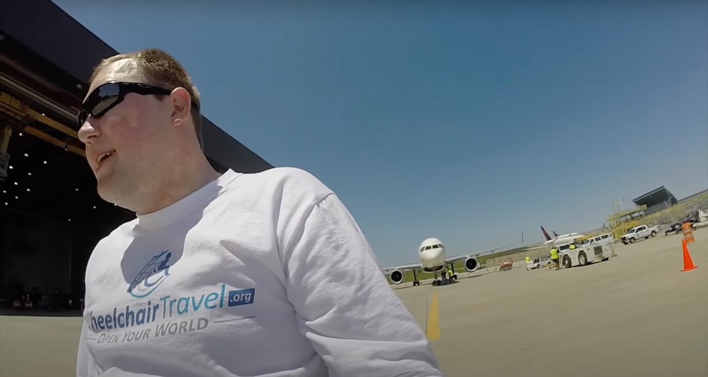 John Morris wearing a white Wheelchair Travel logo t-shirt in front of an airplane.