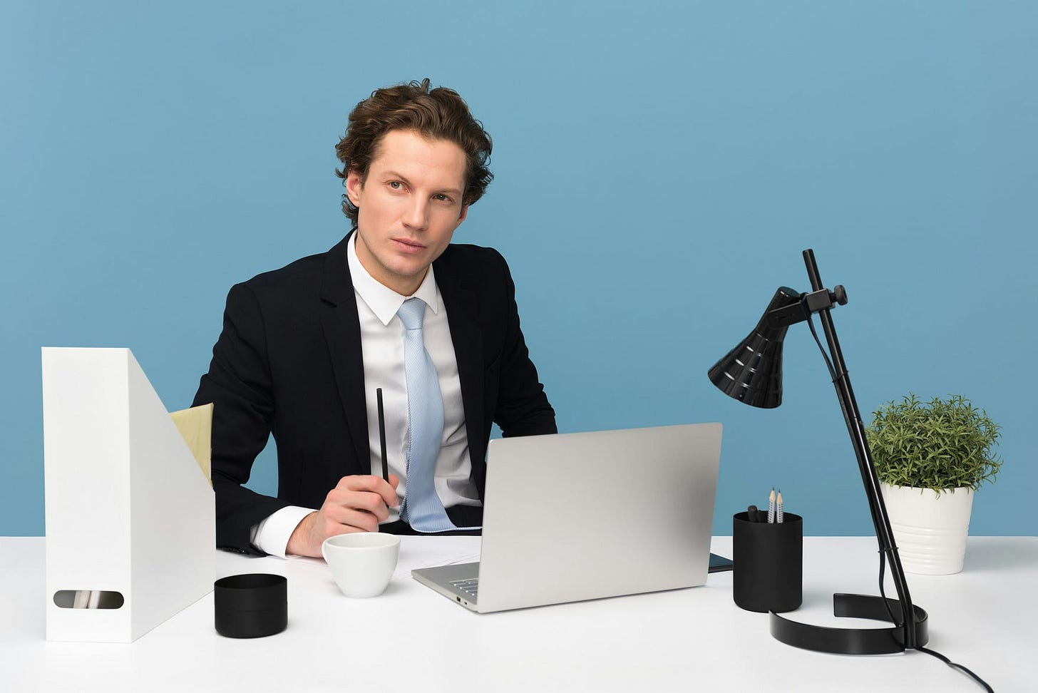 a man in a business suit sitting on his desk and a blue wall behind him a man in a business suit sitting on his desk and a blue wall behind him