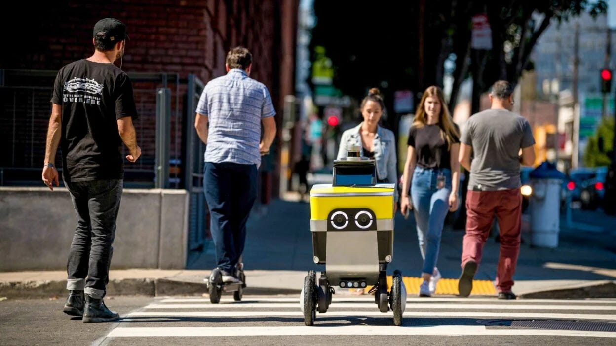 Foto: Serve RoboticsFoto de robô de 4 rodas com formato que lembra um carrinho de sorvetes atravessando faixa de pedestres de uma rua movimentada. No primeiro plano há a pessoas atravessando a pé e em patinetes. No fundo desfocado há sinais de trânsito e arranha céus.