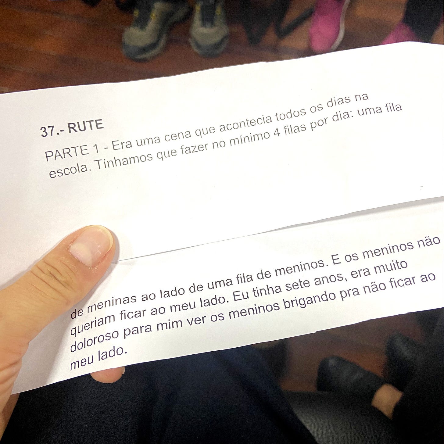 A imagem mostra a minha mão segurando dois pedaços de papel com uma história sequencial escrita. No topo da página há um título que diz "37. RUTE PARTE 1 - Era uma cena que acontecia todos os dias na escola. Tínhamos que fazer no mínimo 4 filas por dia: uma fila de meninas ao lado de uma fila de meninos. E os meninos não queriam ficar ao meu lado. Eu tinha sete anos, era muito doloroso para mim ver os meninos brigando para não ficar ao meu lado”