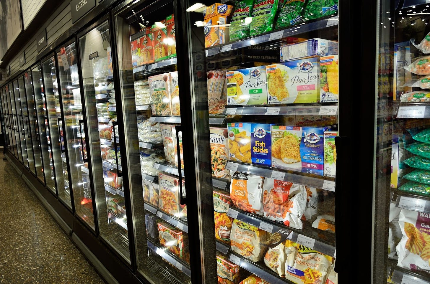a close up of frozen food items in boxes in the grocery store aisle includes a blue box of fish sticks, sweet potato fries, peas and pizza