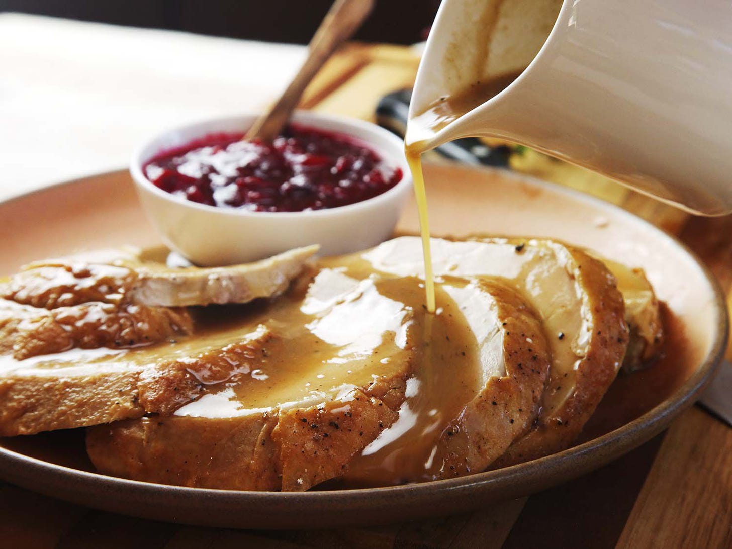 Pouring gravy from a pitcher over a platter of sliced roasted turkey breast meat, with a bowl of cranberry sauce in the background.