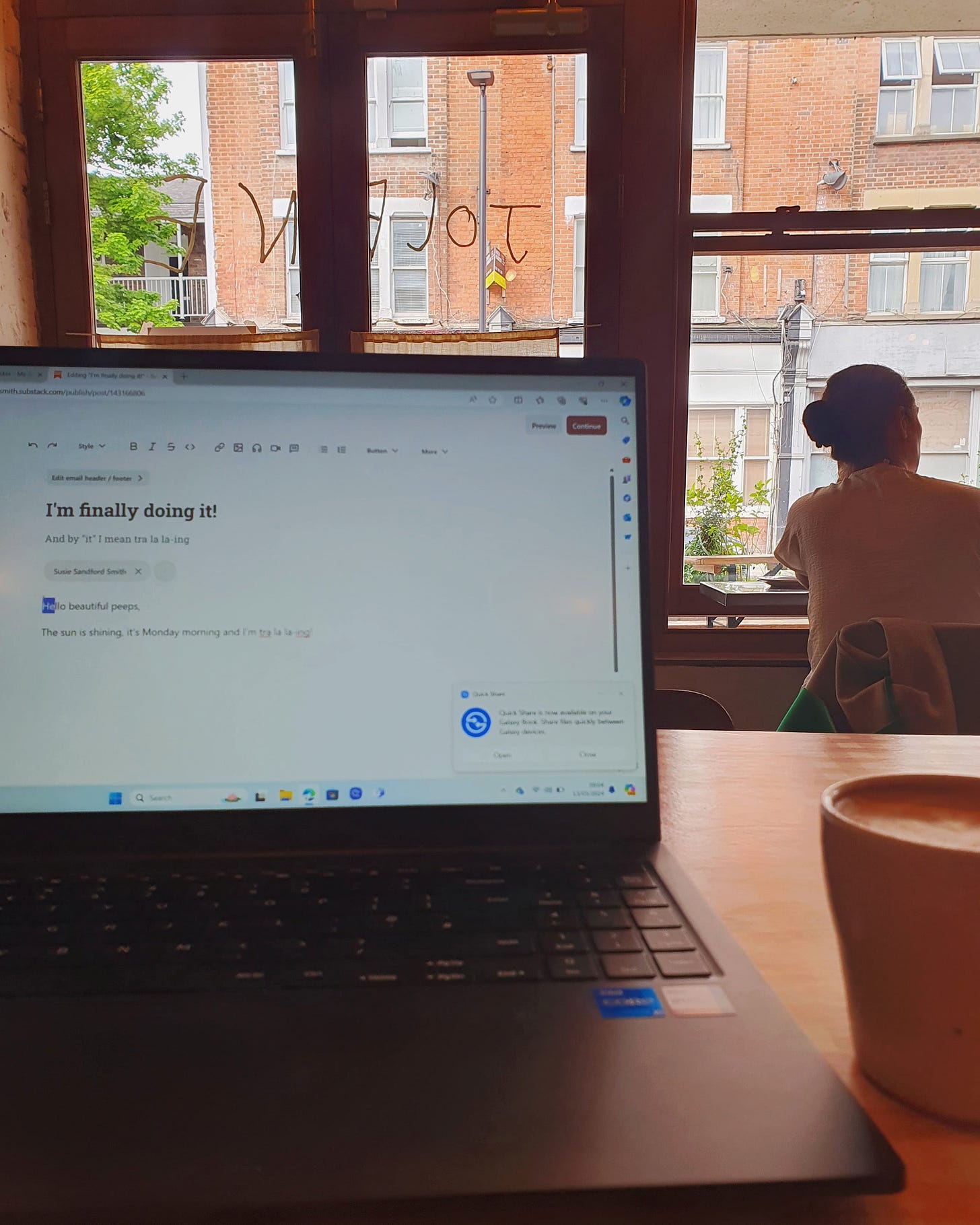 An open laptop with some writing on the screen looking out from a table in a coffee shop.