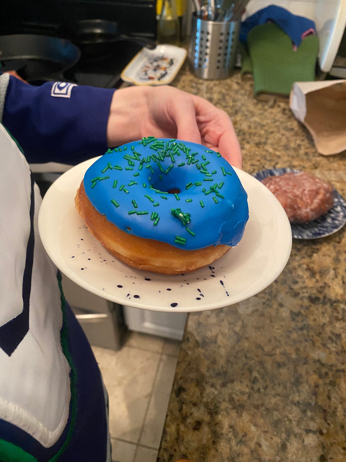 My hand is holding a blue iced donut with green sprinkles. You can see a sliver of my Canucks jersey on the left side, the skate-in-rink design. In the background on another plate is an apple fritter.