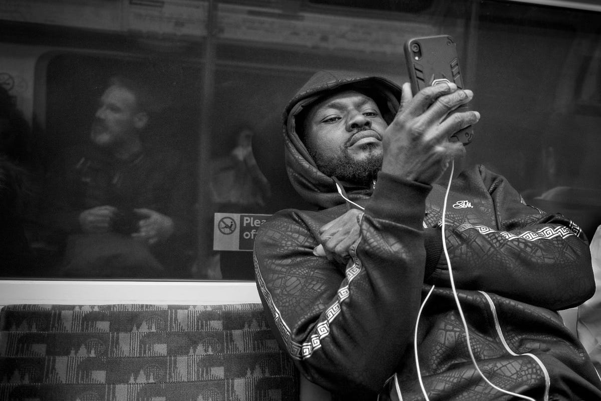 Image of me reflected in a window taking a photograph of a man on his phone on the tube in London.